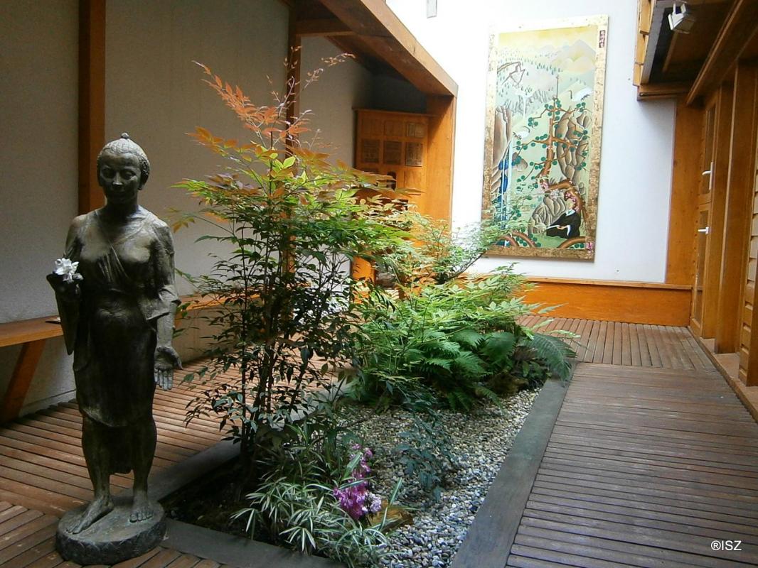 Un jardin japonais serein, avec une statue de Bouddha en pierre, des fougères verdoyantes, une passerelle en bois et une peinture japonaise traditionnelle sur le mur.